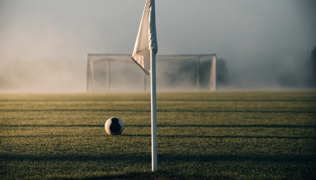 Eine Eckfahne steht am Morgen auf nassem Rasen, Tautropfen hängen im Gras und über dem Platz liegt leichter Dunst.