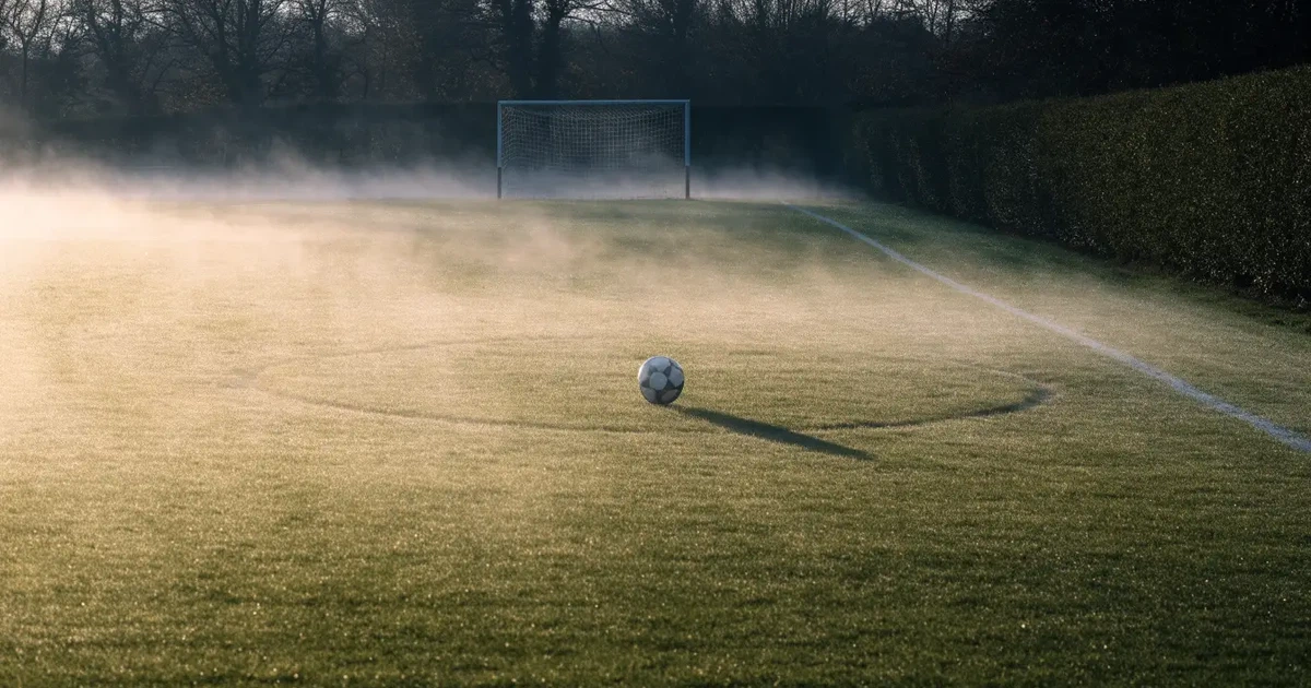 Ein Fussballplatz im Morgennebel, im Mittelkreis liegt ein einzelner Ball und der Rasen glänzt vom Tau.