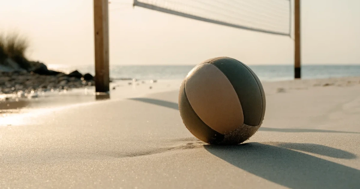 Ein Volleyball liegt auf feinem hellem Sand, ein gespanntes Netz verschwimmt dahinter, das Licht kommt von einem weiten Meer.