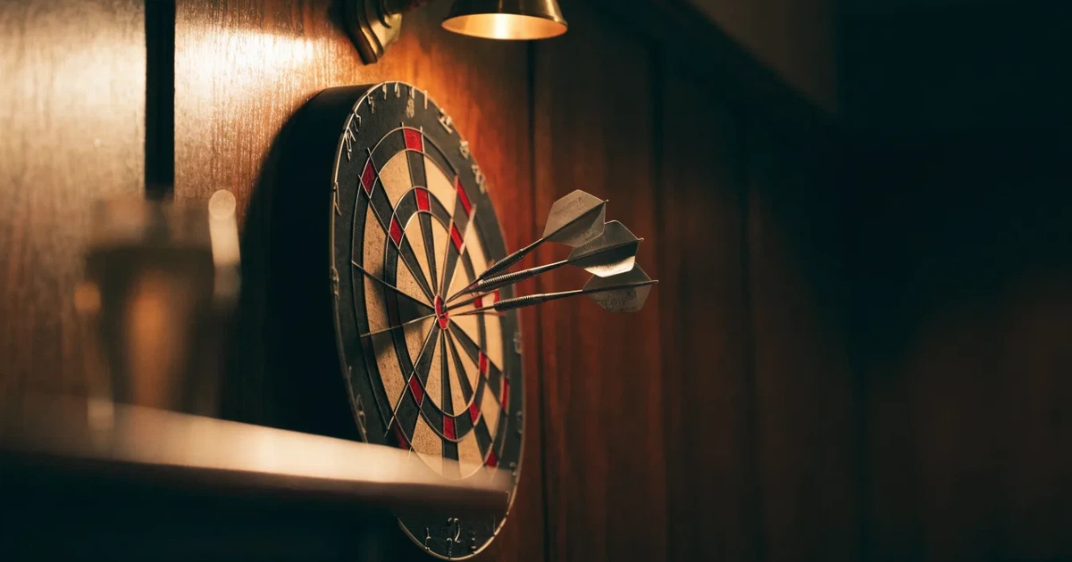 Drei Dartpfeile stecken in einer Dartscheibe an einer Holzwand in einer dämmrigen Kneipe, warmes Lampenlicht fällt von oben
