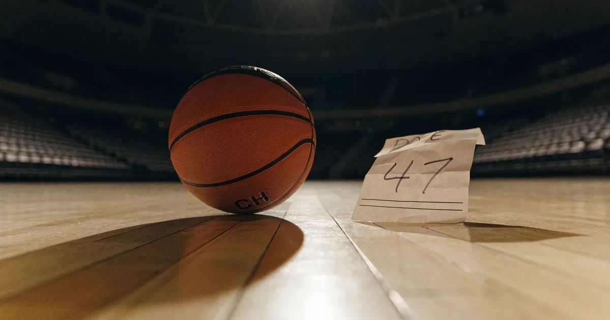 Ein Basketball liegt auf dem hellen Parkett einer leeren Halle, im Hintergrund steigen die Tribünenreihen im Halbdunkel an.