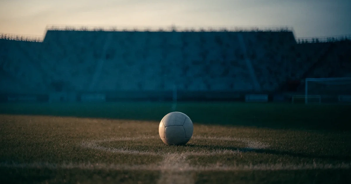 Ein Fussball liegt auf dem Mittelpunkt eines leeren Rasens, im Hintergrund steigen steile Tribünen in der Dämmerung auf.