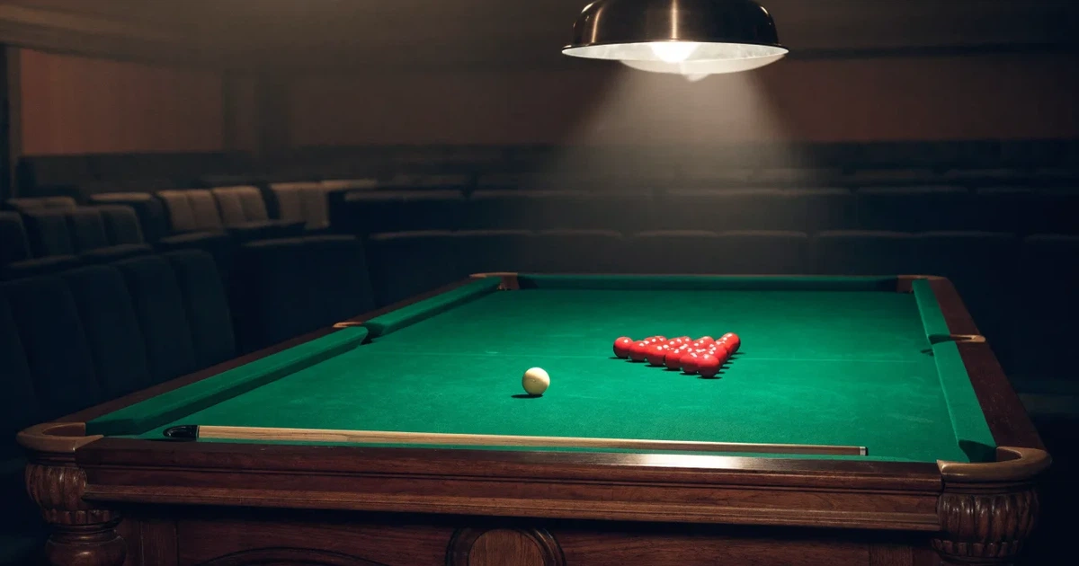 Ein Snookertisch mit grünem Tuch und einem Dreieck aus roten Kugeln unter einer tief hängenden Lampe in einer leeren Halle.