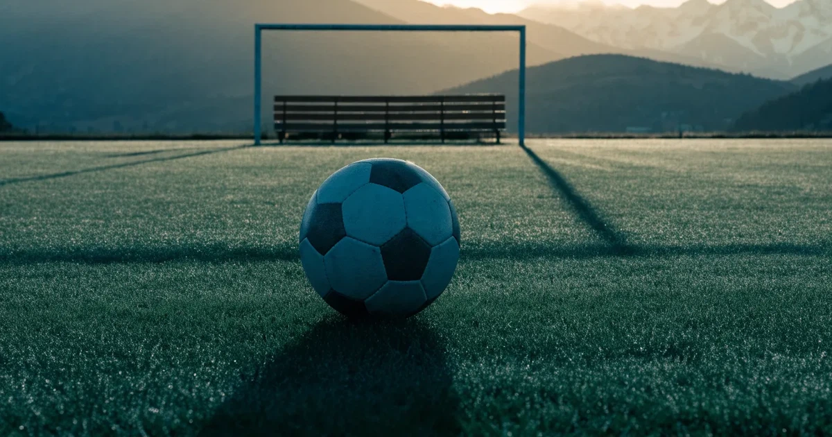 Ein Fussball liegt am frühen Morgen auf dem Rasen eines leeren Fussballfeldes, dahinter eine kleine Tribüne und schneebedeckte Berge.