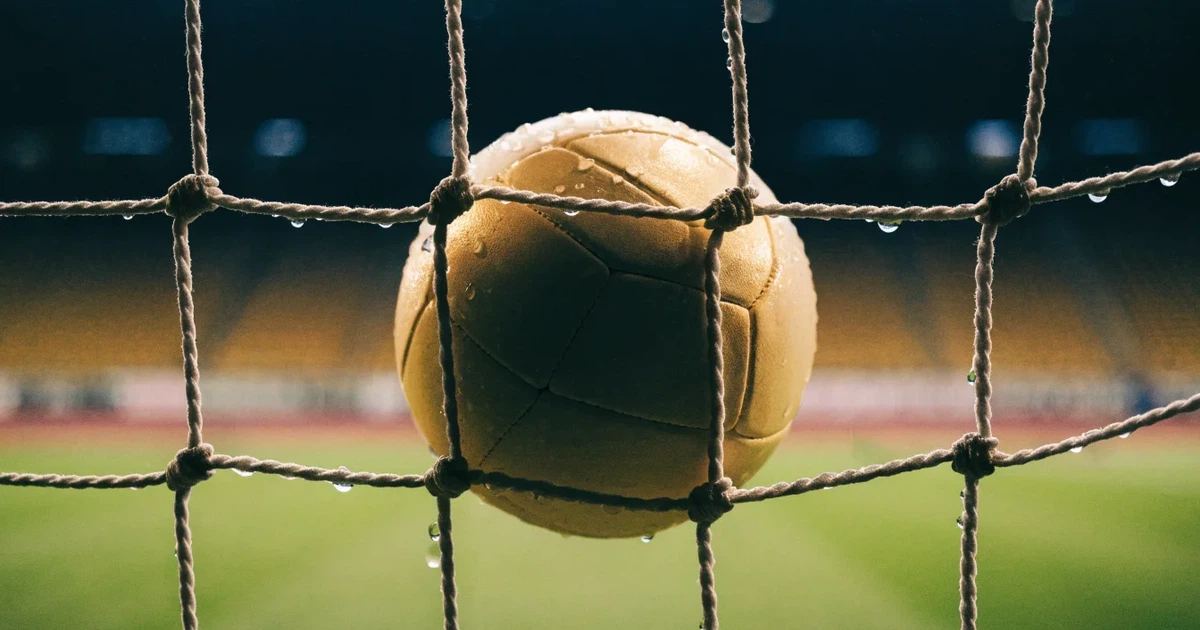 Ein Fussballnetz in Nahaufnahme, in dem ein Ball hängt, Wassertropfen glänzen auf den Maschen.