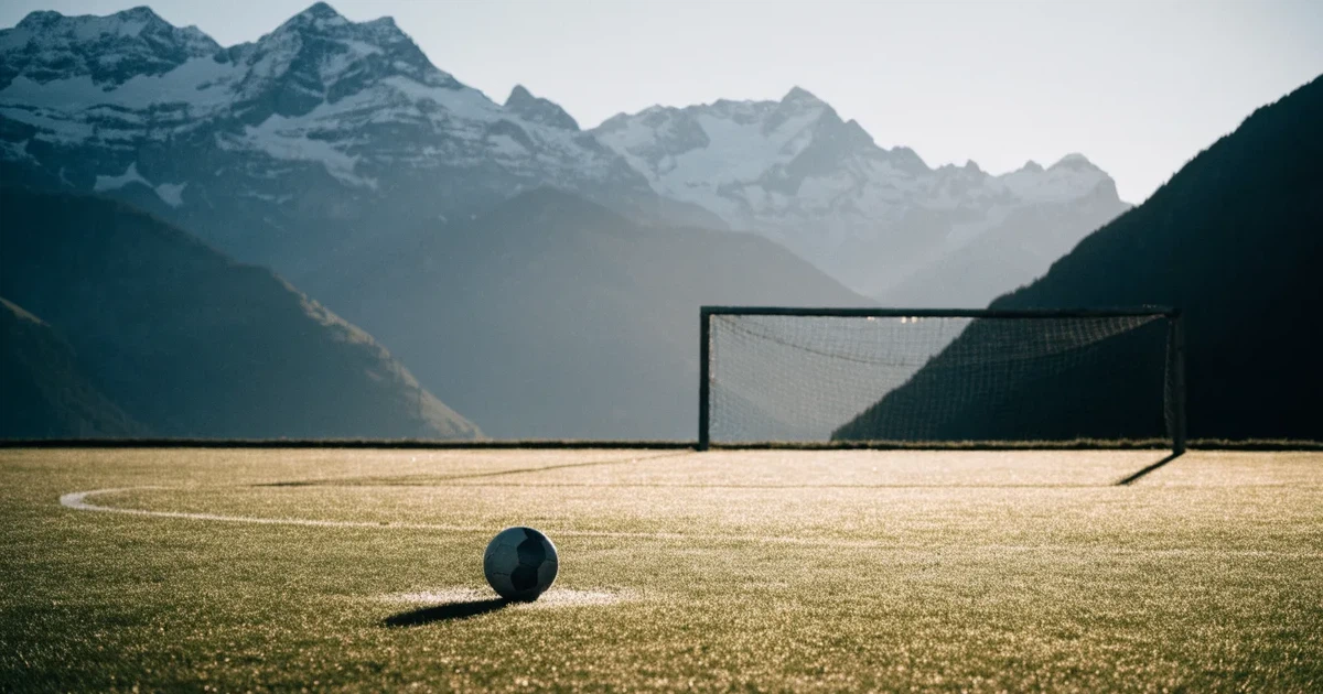 Ein Dorf-Sportplatz mit einem Ball am Strafraumrand, dahinter schneebedeckte Alpengipfel im klaren Morgenlicht